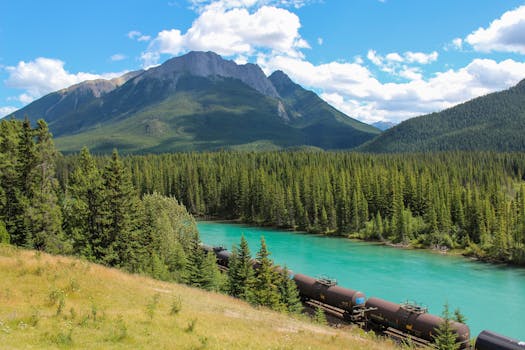 Train travels through lush forest with mountains and turquoise river under a sunny sky.