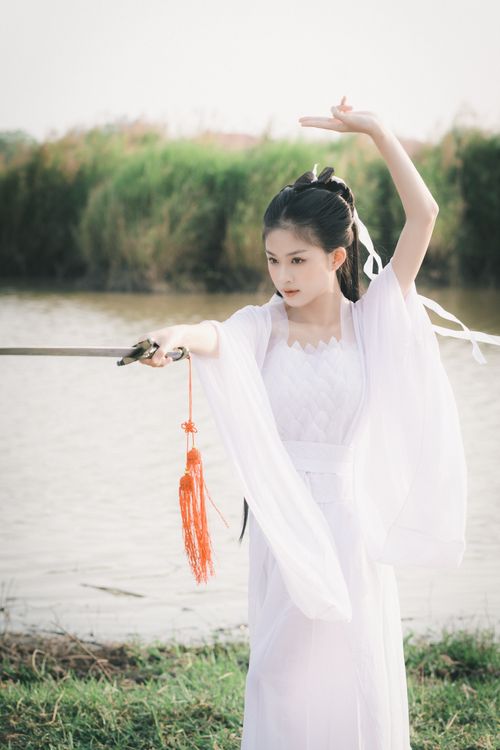 Free Young Asian woman in a white dress elegantly posing with a sword by the water. Stock Photo