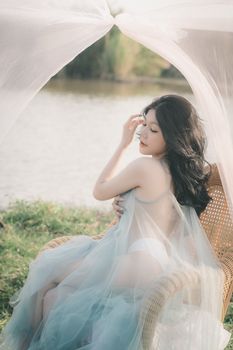 A young Asian woman poses gracefully in a sheer dress with tulle by the lakeside on a sunny day.