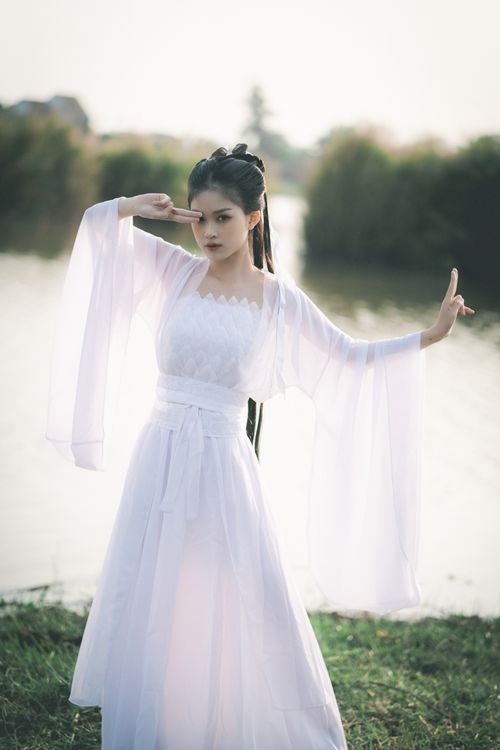 Free A woman in an elegant white dress posing gracefully by a peaceful lakeside. Stock Photo