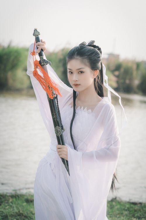 Free Elegant young woman in white traditional dress holding a sword by a river. Stock Photo