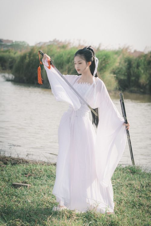 Free A young woman in a white traditional dress holding a sword by the water, embodying elegance and strength. Stock Photo