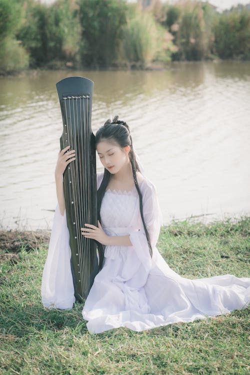 Brunette Girl Wearing a White Dress Kneeling with a String Instrument by a River