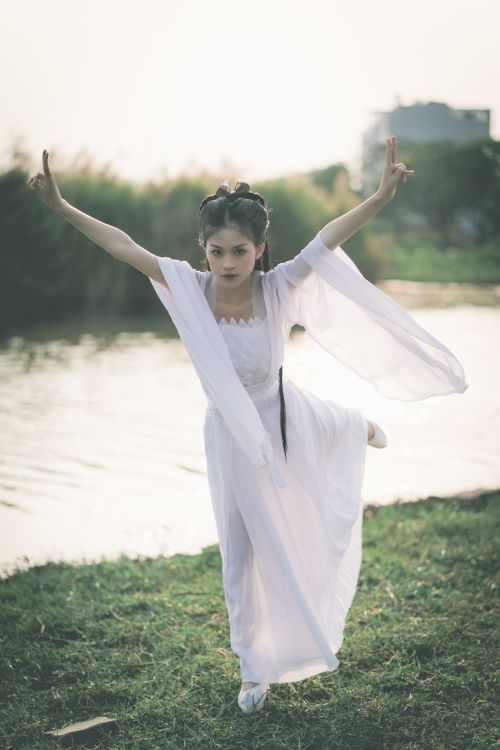 Free A young woman in a flowing white dress poses gracefully by a riverside, embodying elegance and balance. Stock Photo