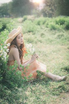 A young woman with long hair in a summer dress and hat sitting in a grassy field holding flowers.