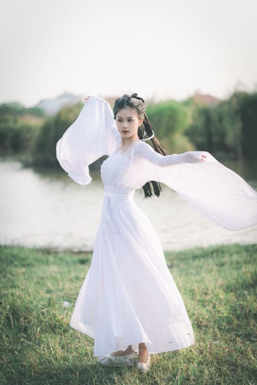 Free Portrait of a young woman in a white dress with arms raised near a lakeshore, embodying serene elegance. Stock Photo