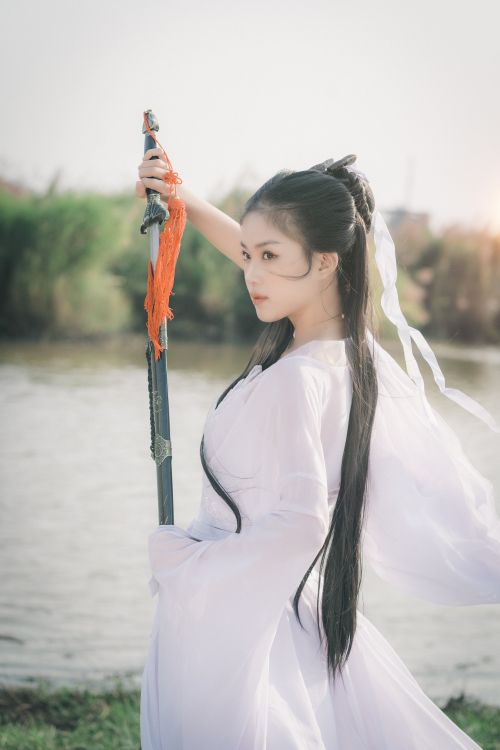 Free Beautiful East Asian woman poses gracefully with a sword, embodying elegance and tradition by the river. Stock Photo