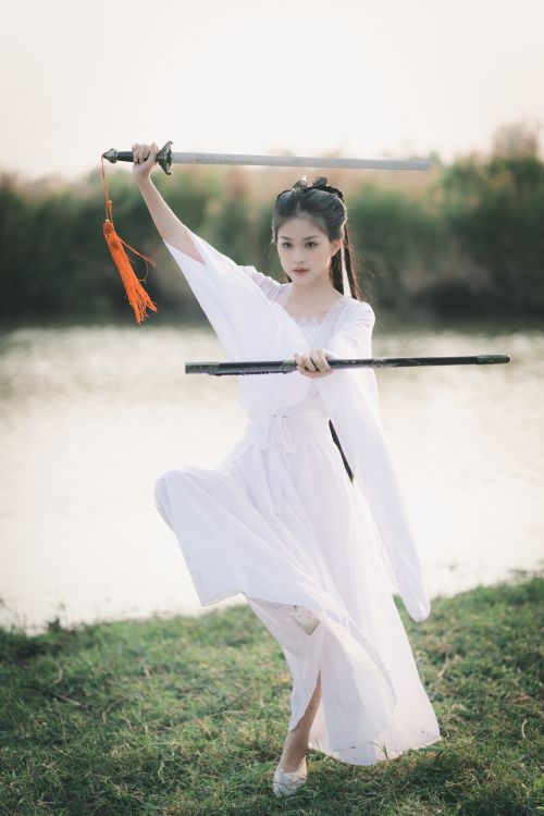 Free Asian woman practicing Tai Chi sword kata in a white dress at a serene park setting. Stock Photo
