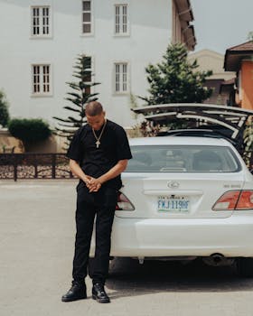 Man in black t-shirt posing beside a white car on a sunny city street.