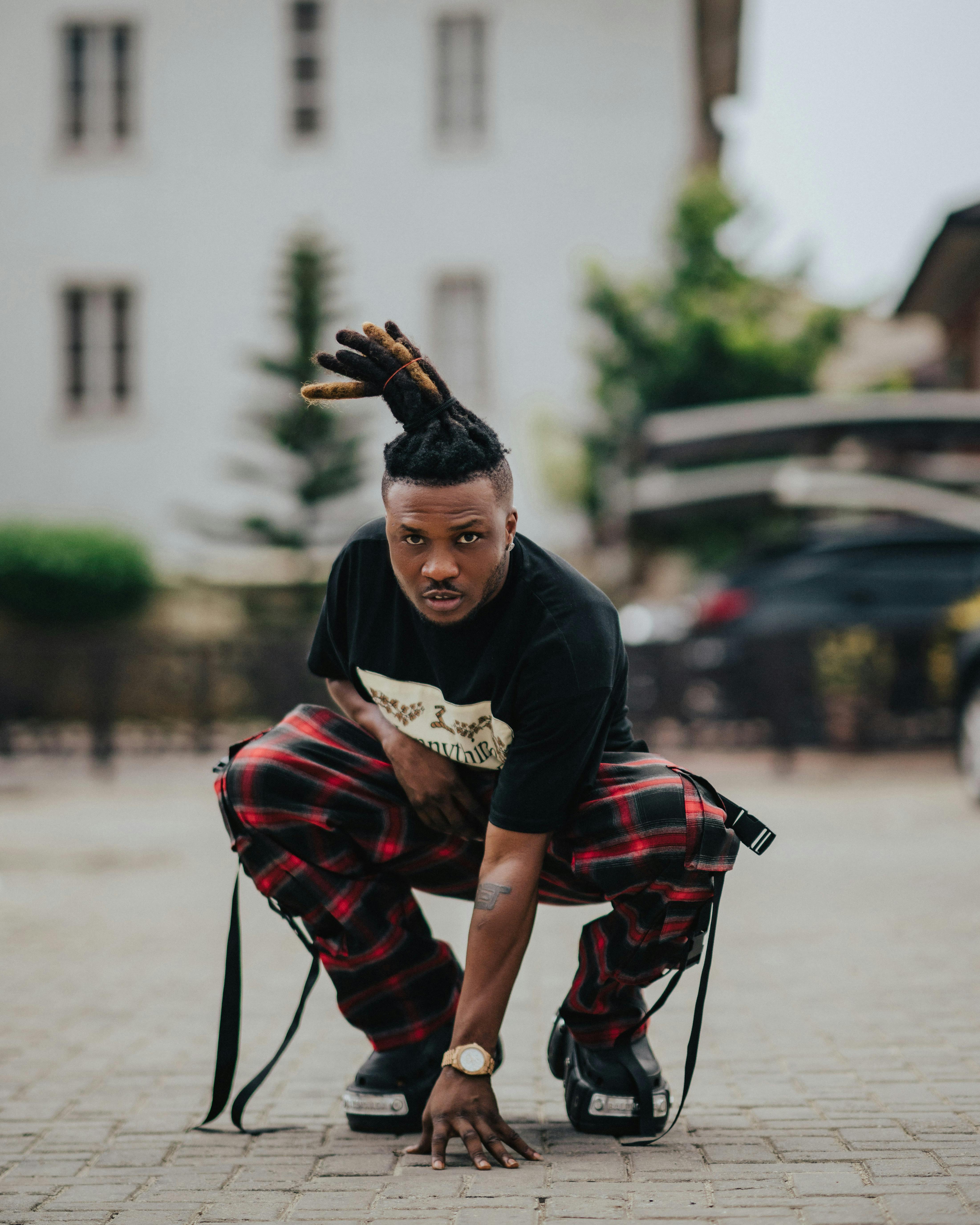 Young man crouching on city street in hip hop style with bold dreadlocks and plaid pants.