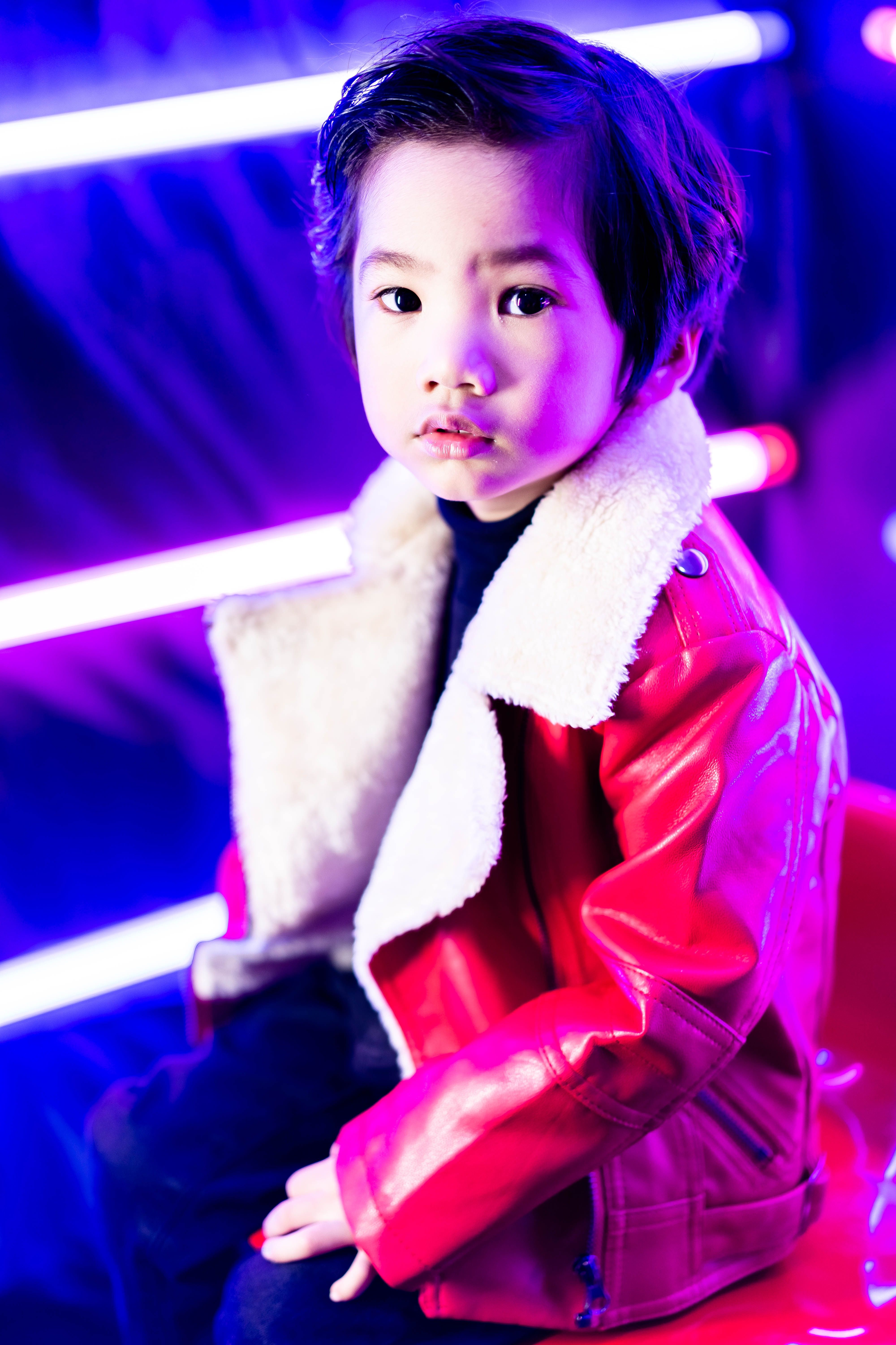 A fashionable young child in a red jacket poses with vibrant neon lighting for a striking portrait.