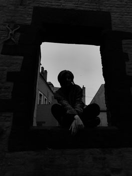 Black and white silhouette of a person sitting cross-legged in a window frame against a cityscape.