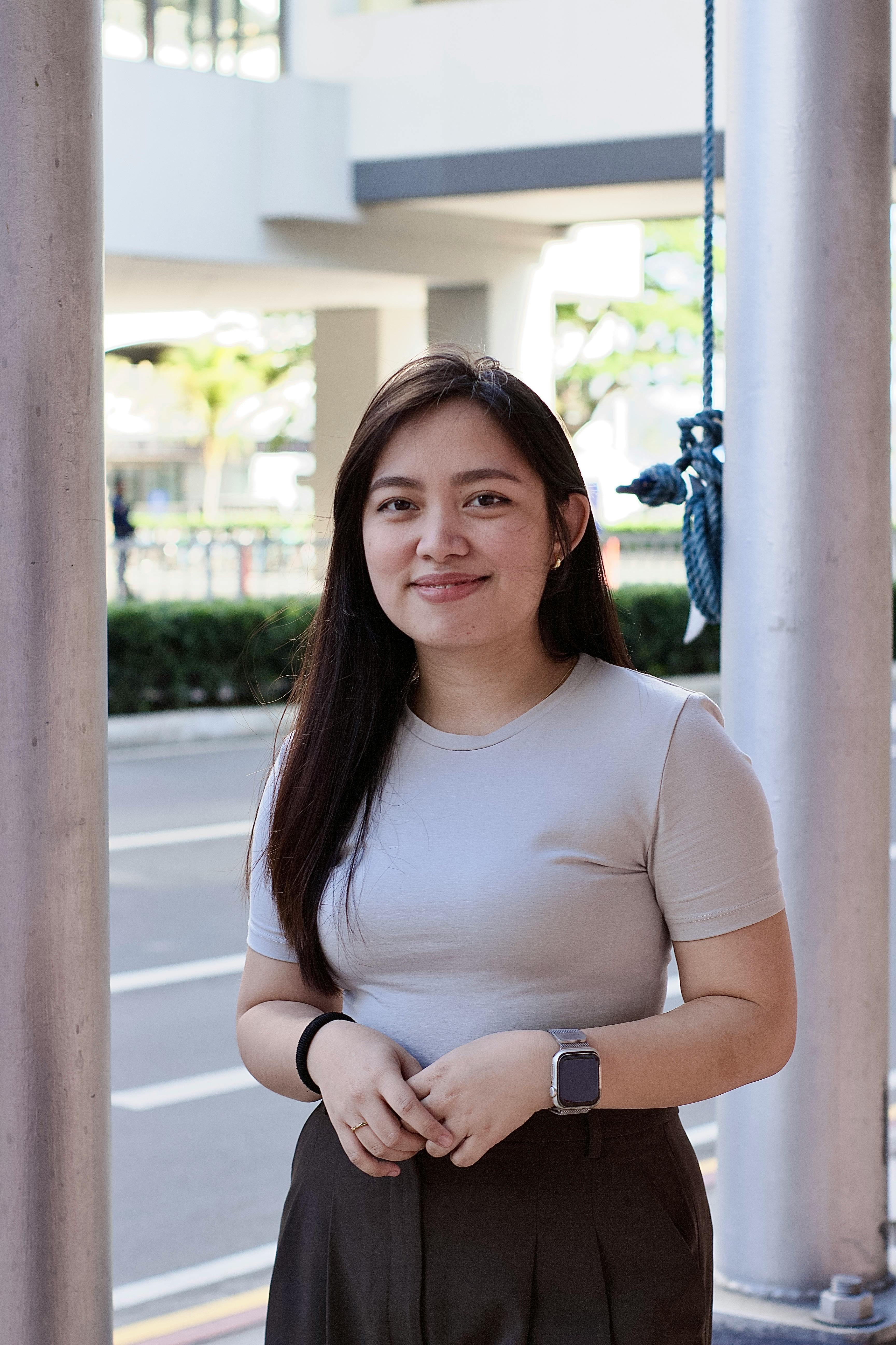 A smiling young woman in casual attire stands outdoors, exuding a warm and approachable vibe.