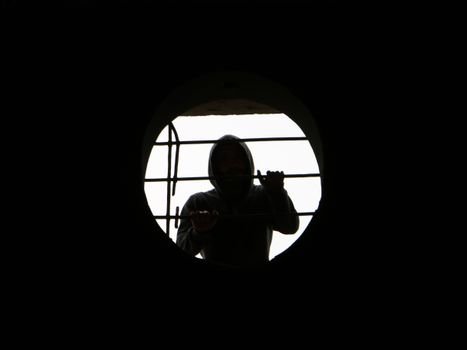 Silhouette of a person in a hoodie behind a round, barred window creating a mysterious scene.