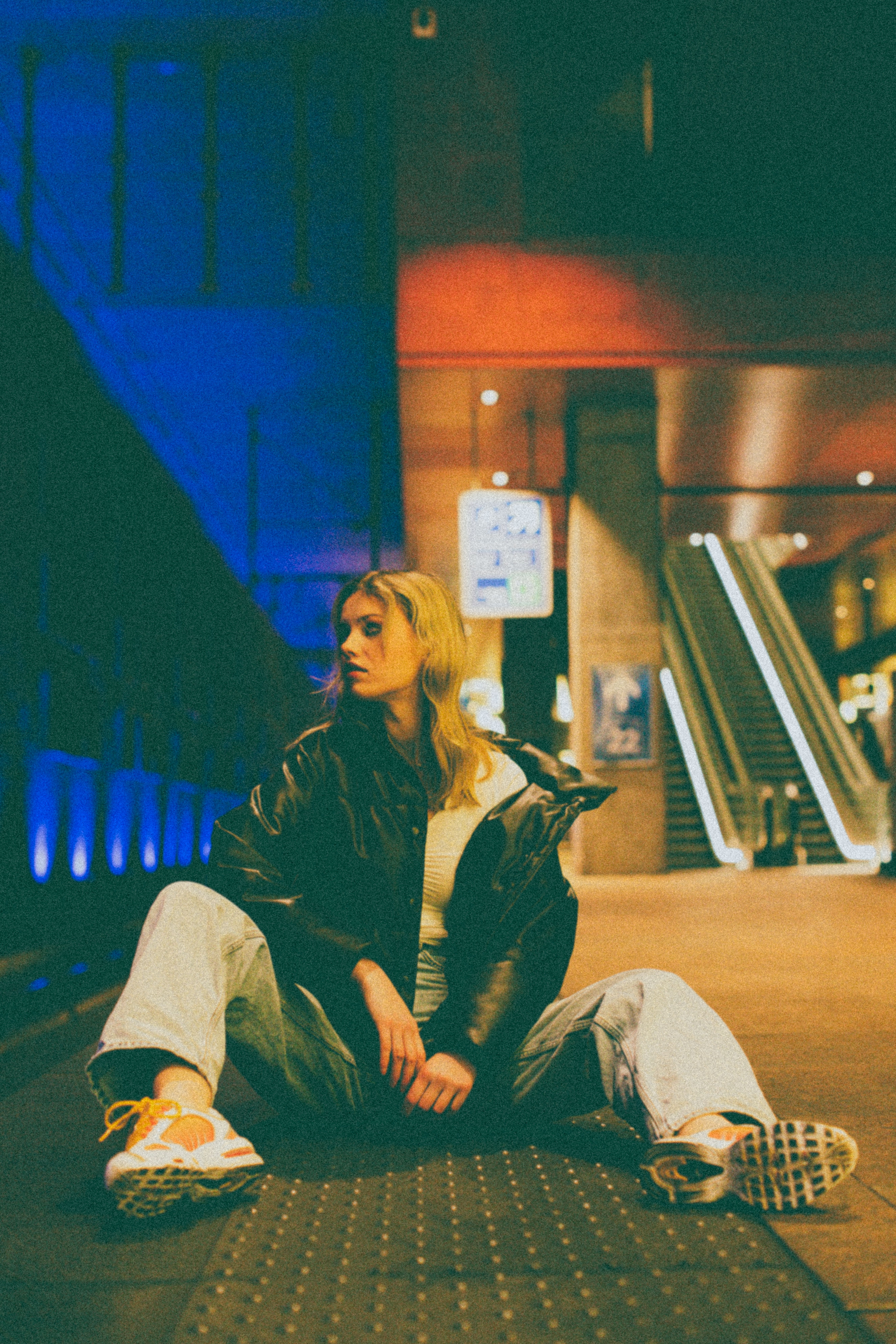 A young woman in casual attire sits stylishly on a subway platform with vibrant lights.