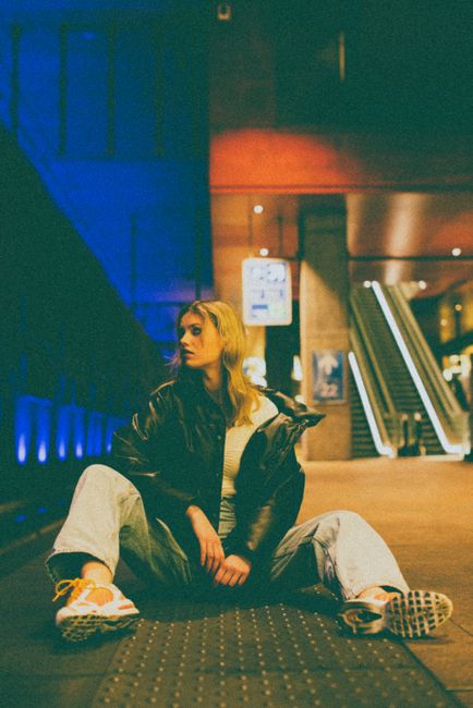 A young woman in casual attire sits stylishly on a subway platform with vibrant lights.
