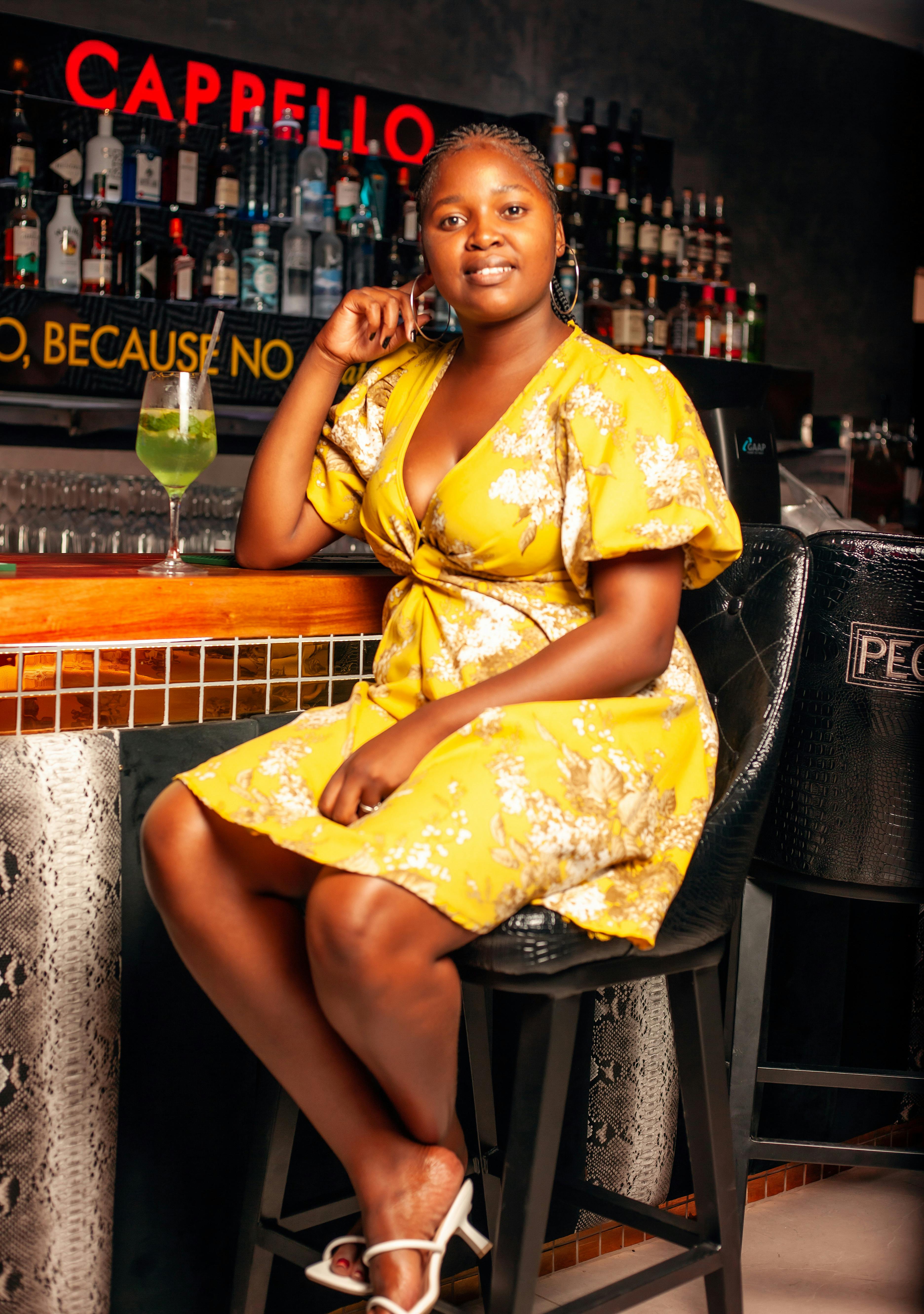 A woman in a yellow dress poses confidently at a bar, surrounded by an array of bottles and soft lighting for a welcoming atmosphere.
