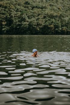 A man wearing a swim cap enjoys a peaceful swim in a serene forest river.