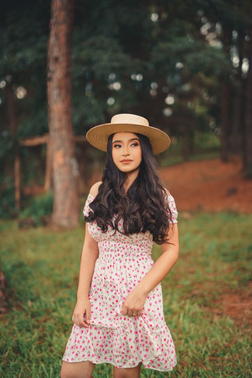 Free Portrait of a young woman wearing a floral dress and sun hat, standing in a forest setting. Stock Photo