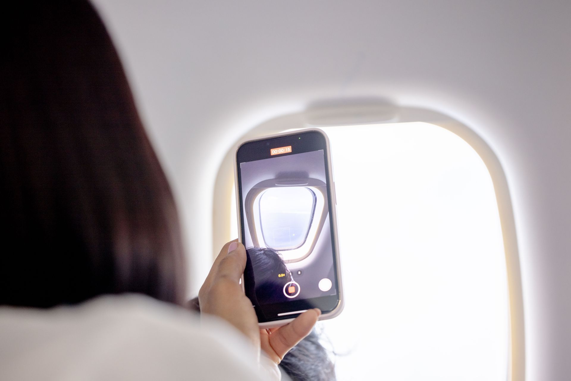A woman records a video of the sky through an airplane window using her smartphone.
