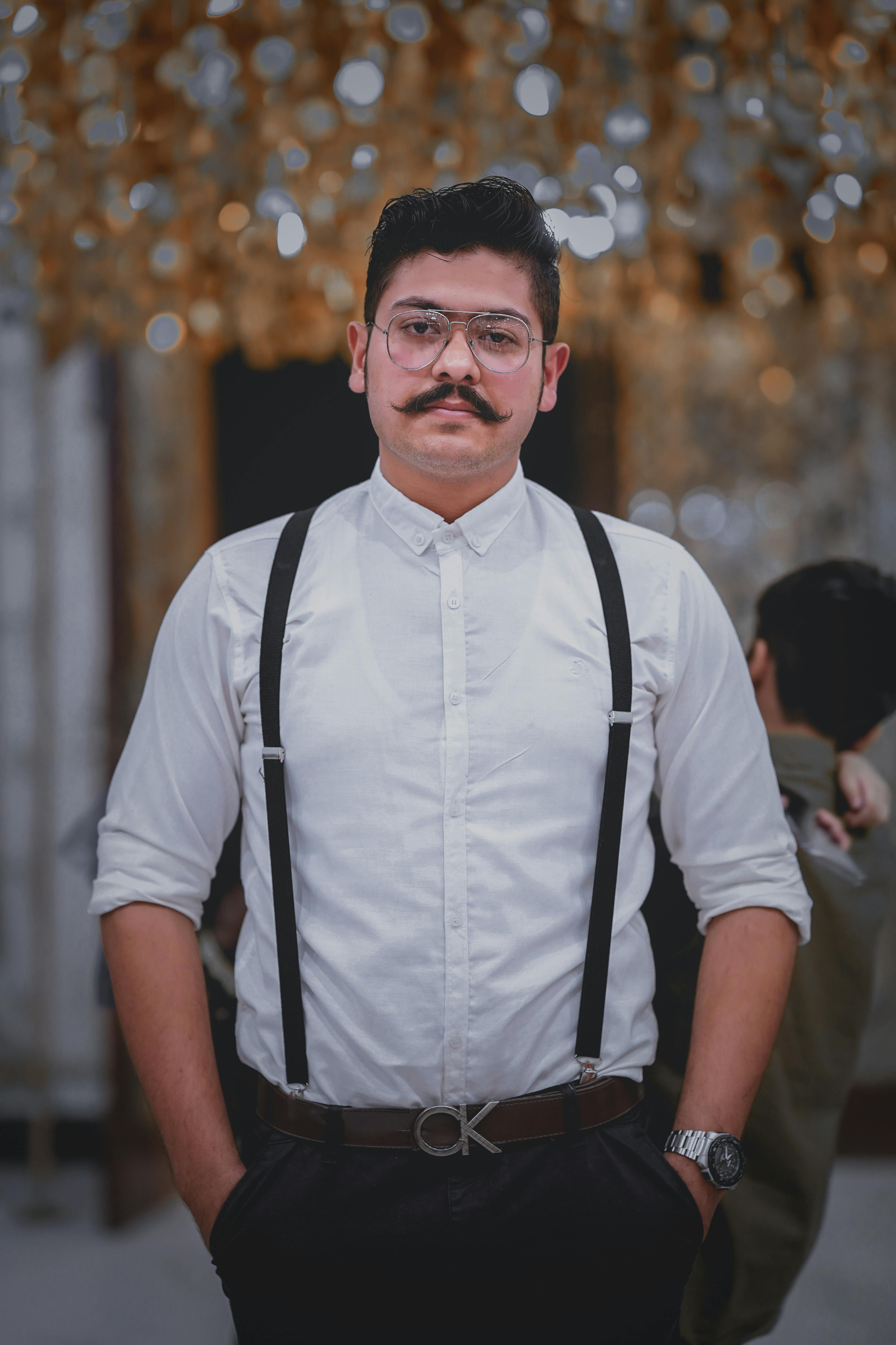 Free Young man with a mustache wearing eyeglasses and suspenders posing stylishly indoors. Stock Photo