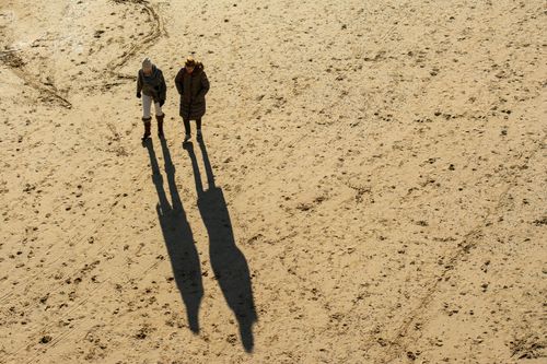Free Two women in warm clothing stroll on a sandy beach casting long shadows. Stock Photo