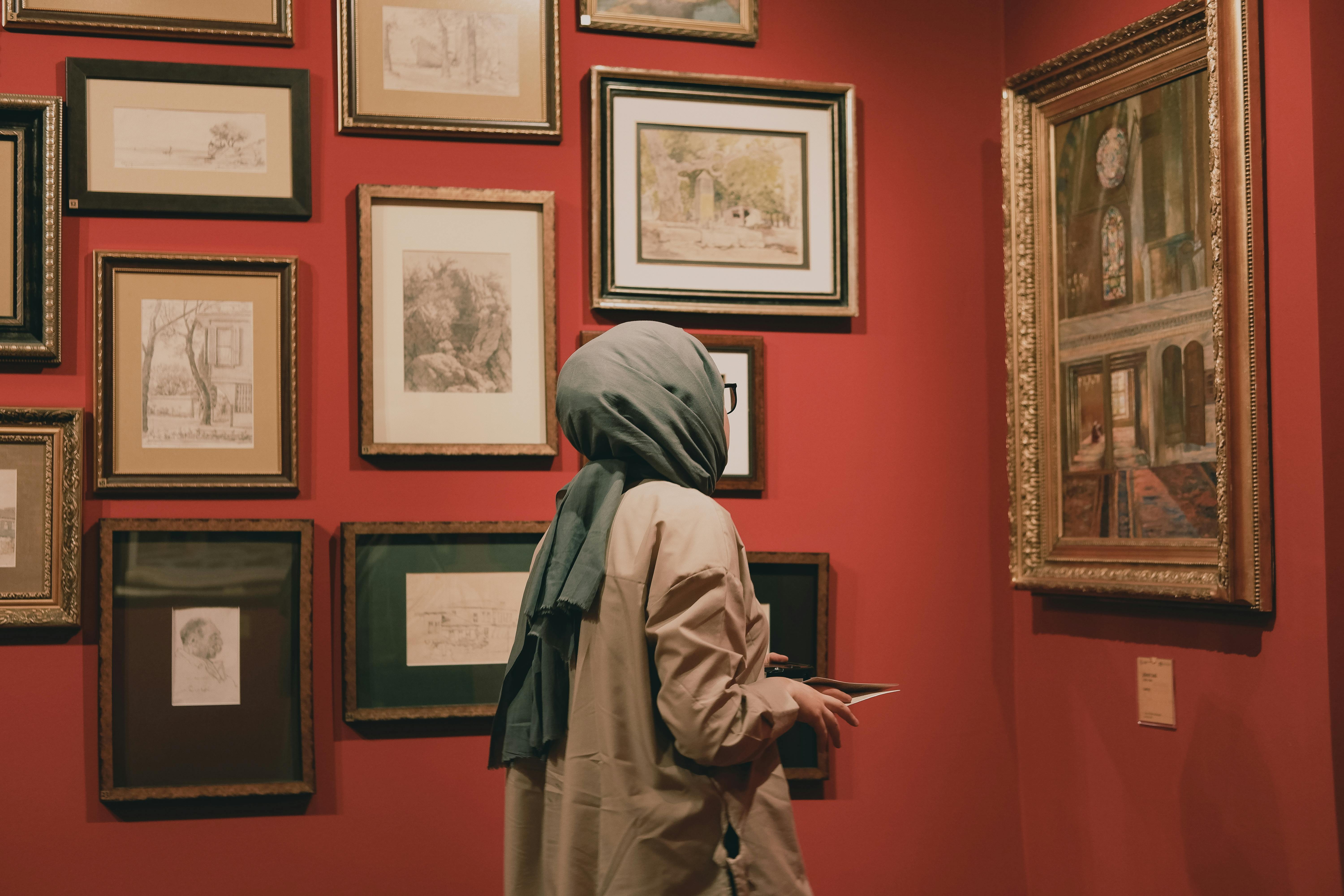 A woman in a hijab observes paintings in a red-walled art gallery.