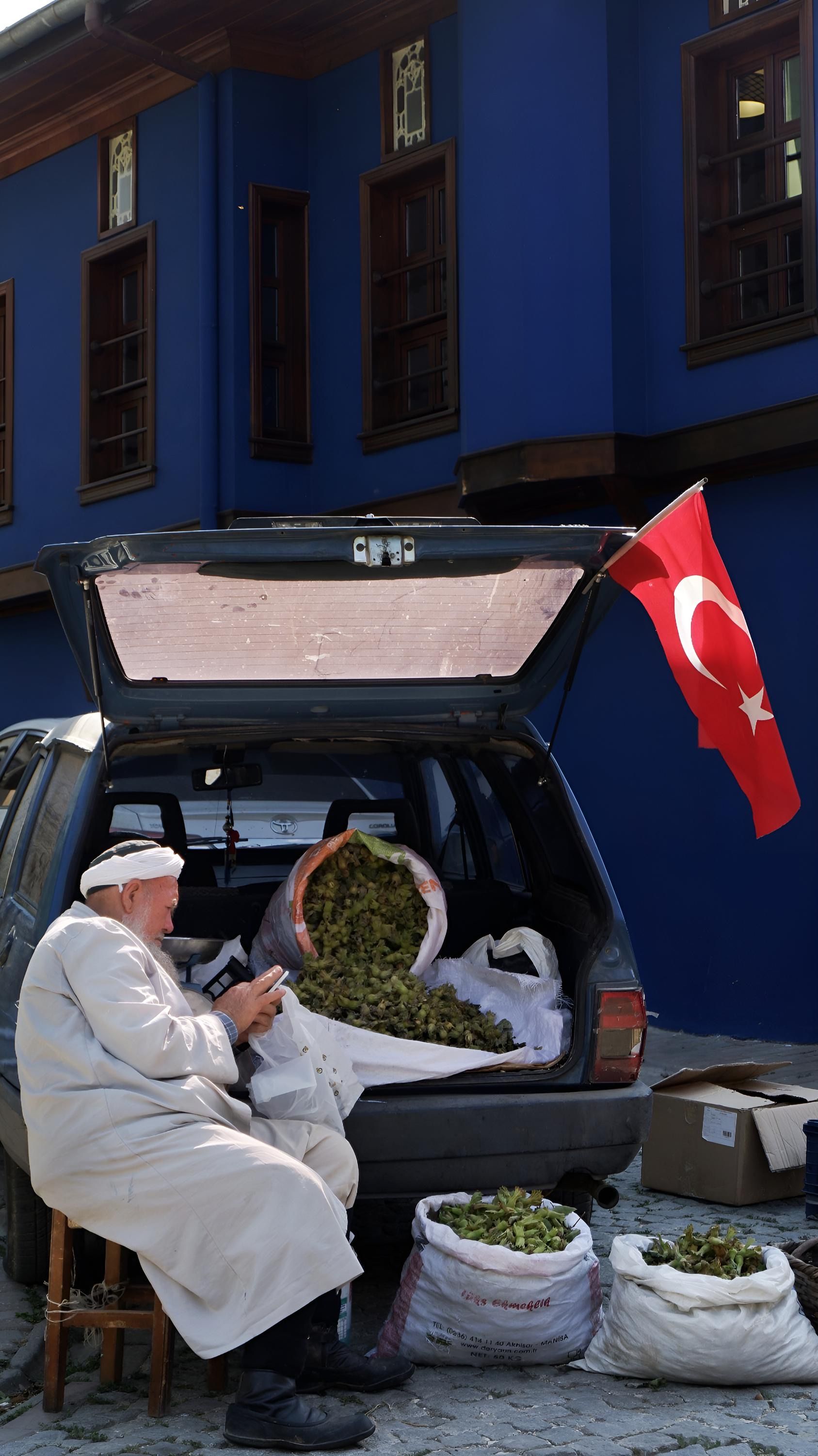 Elderly man sells grapes from his car trunk on a Turkish street with a national flag.