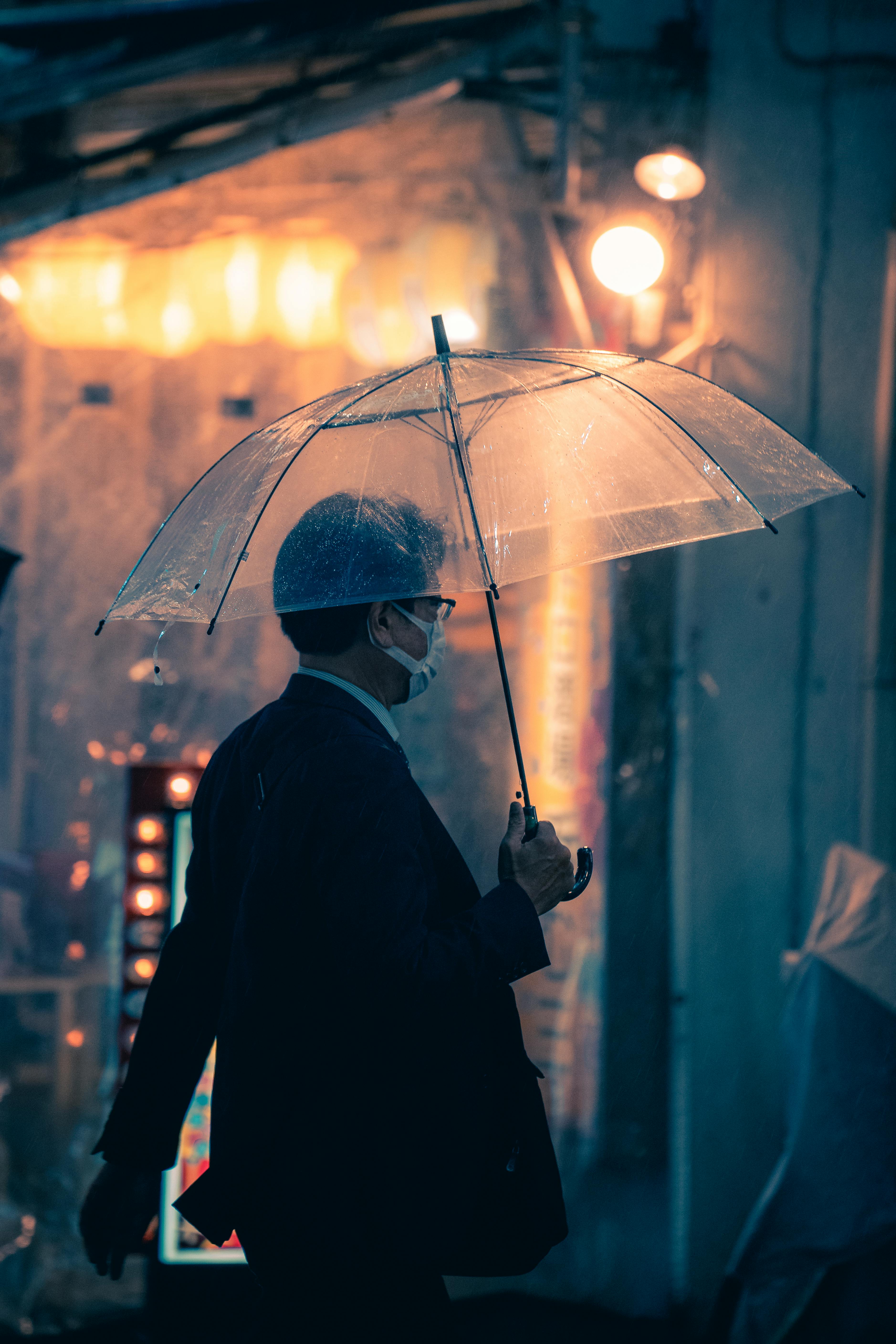 A man wearing a face mask walks under a transparent umbrella on a wet urban street at night.
