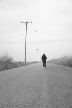 Black and white photo of a person walking along an isolated road with utility poles.