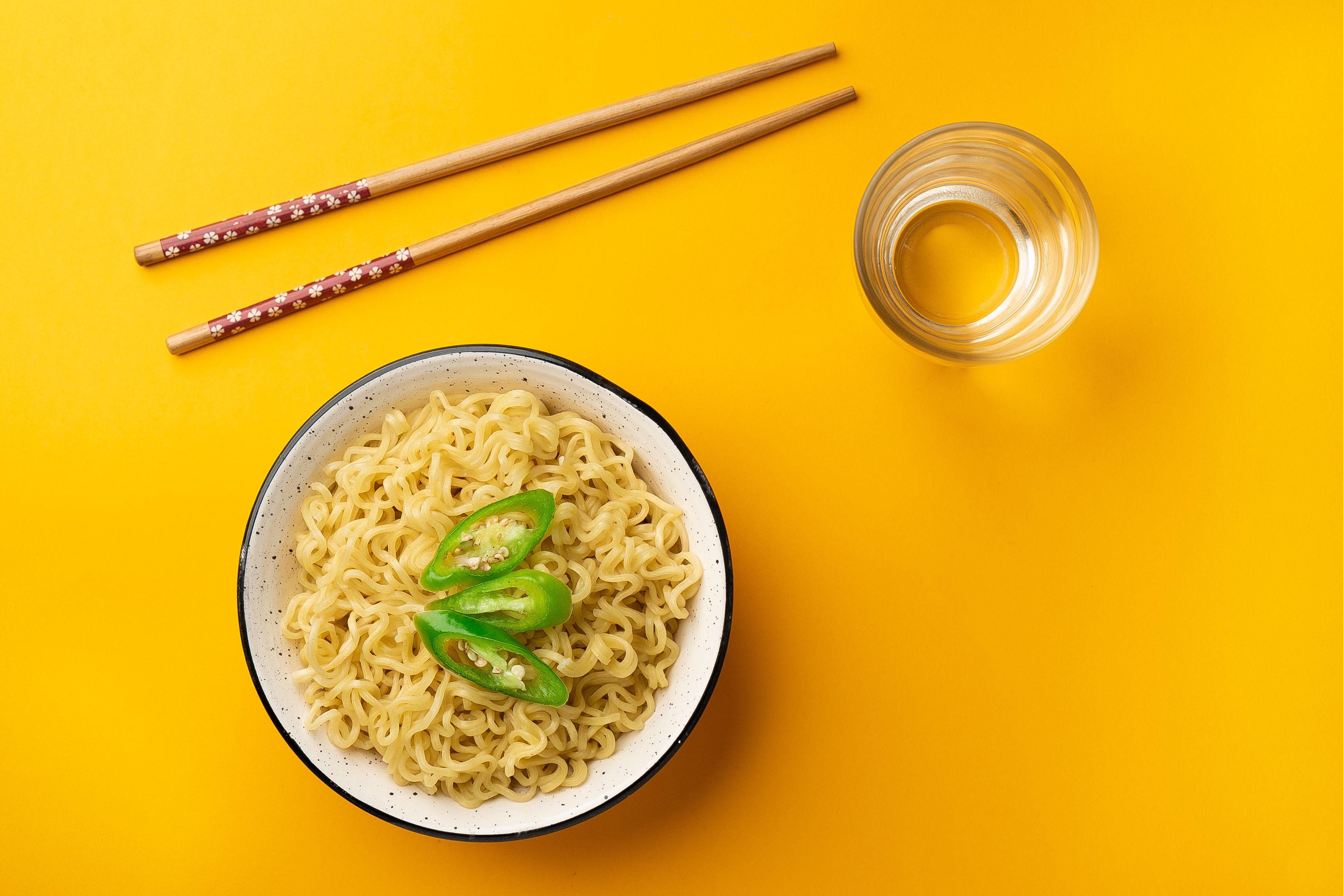 Flat lay of a bowl of noodles with green pepper slices, chopsticks, and a glass of water.
