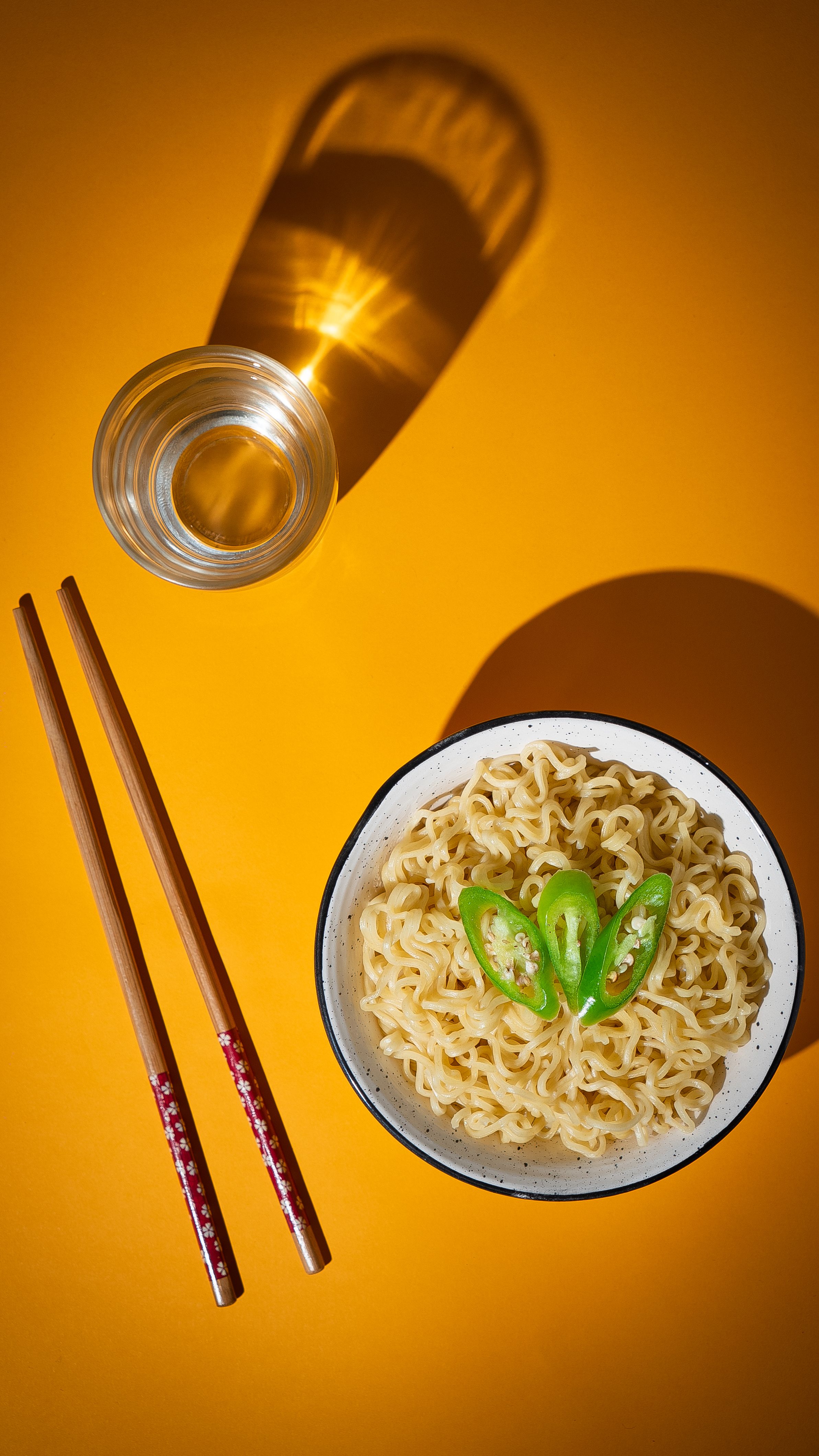 Top view of Asian noodles with green peppers and chopsticks on a striking yellow background.