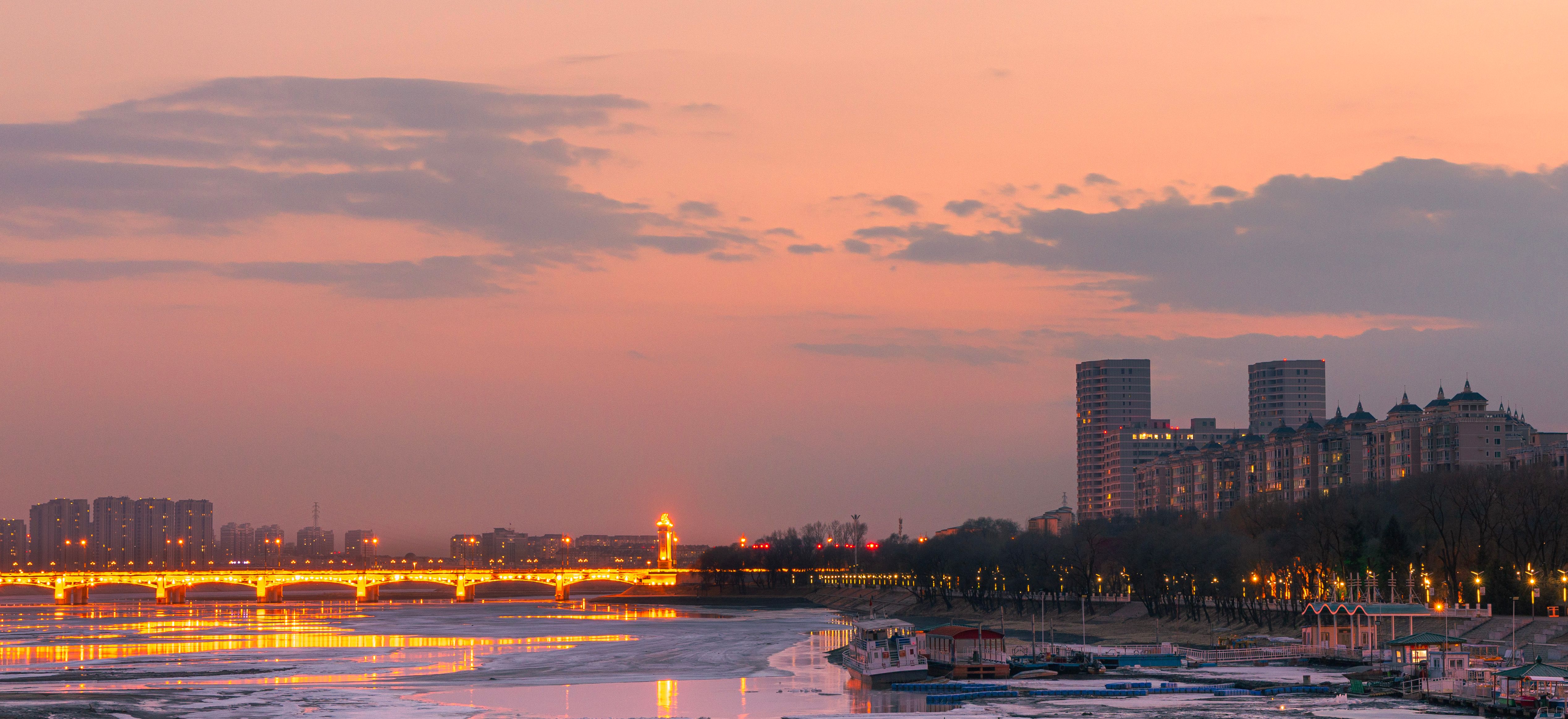 Pemandangan kota dengan jembatan yang diterangi dan bangunan saat senja di atas sungai yang tenang. Via Pexels (by Lianyou)