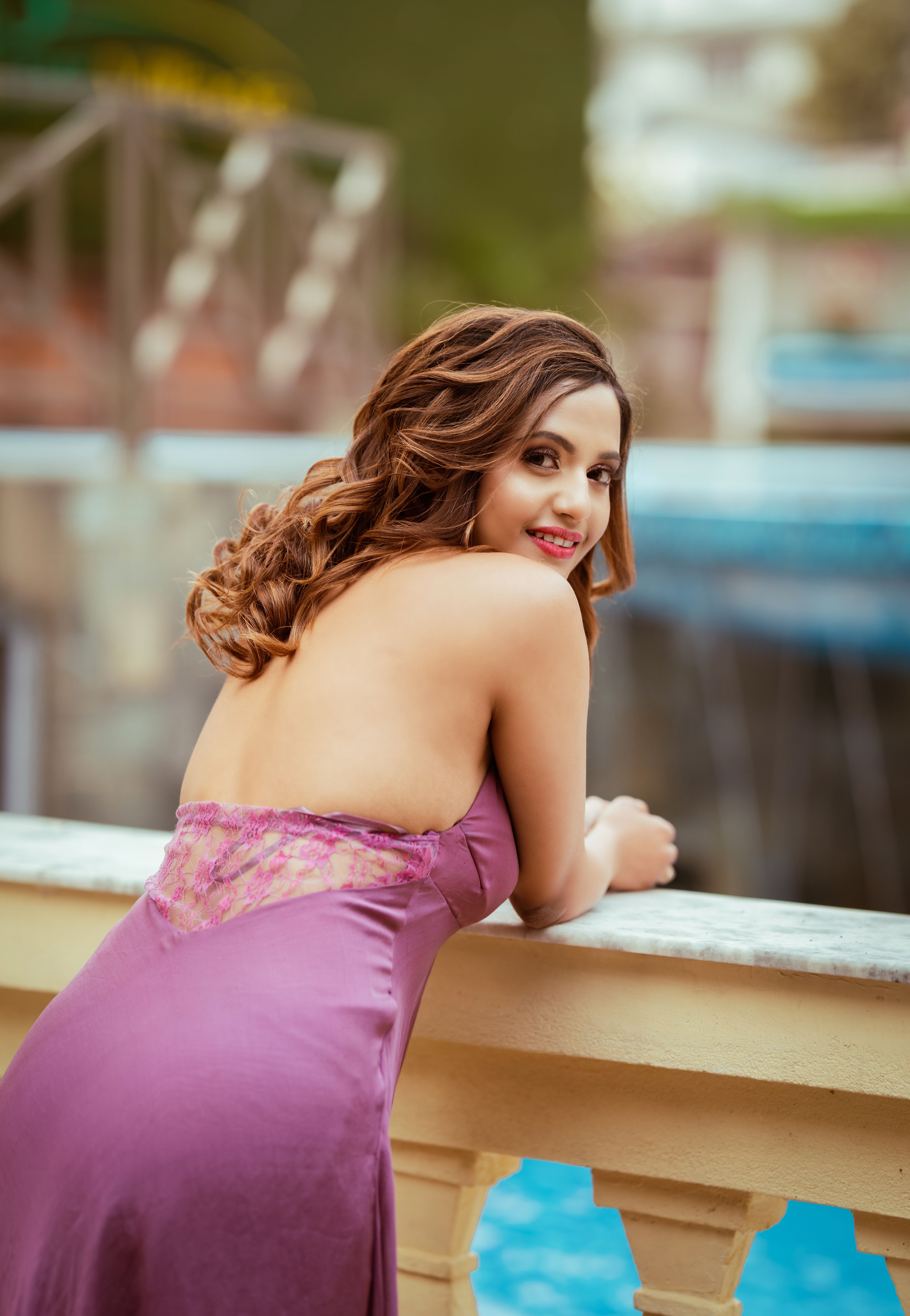 A woman in a purple dress poses elegantly by a poolside balustrade, exuding glamour and charm.