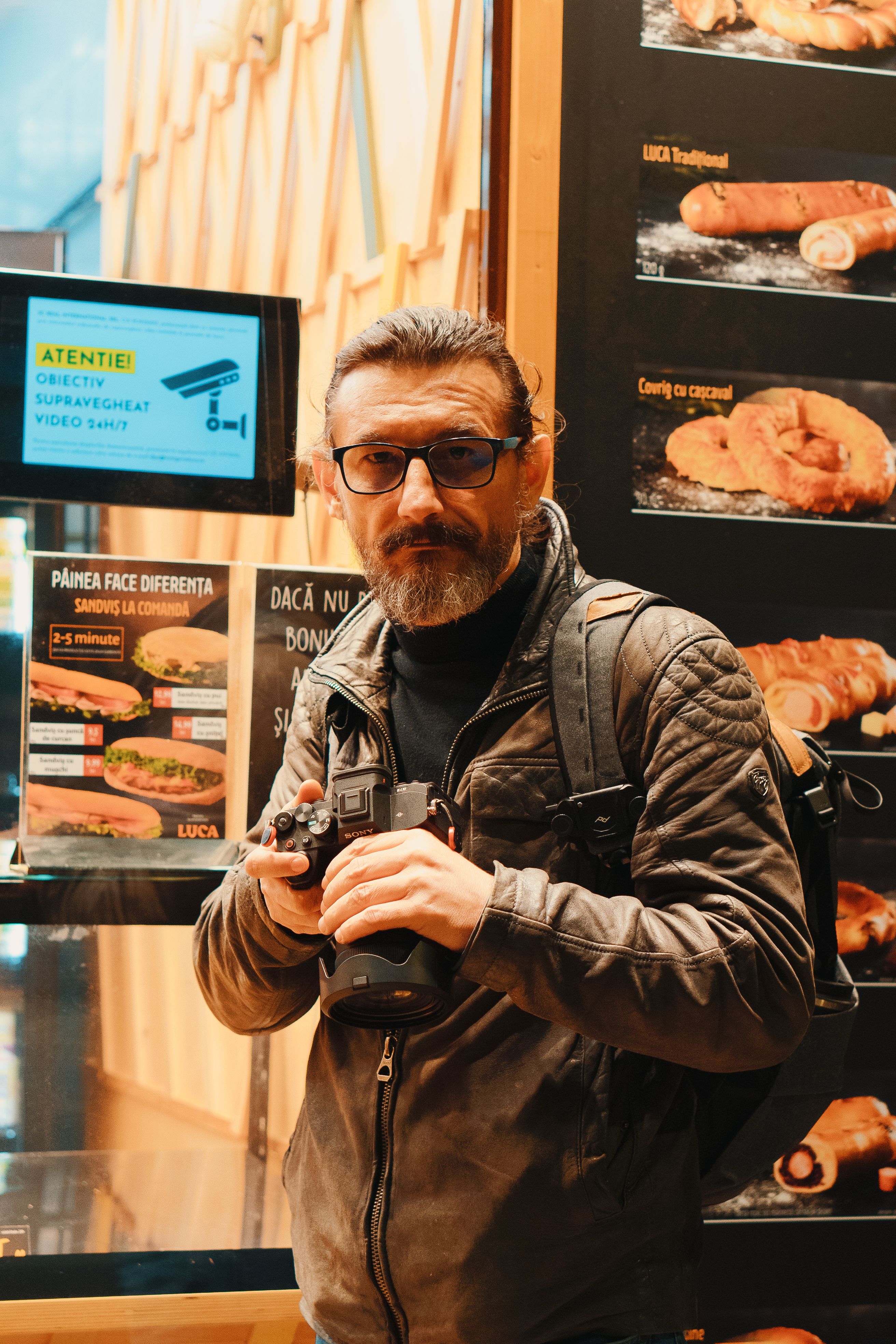 Photographer standing in a fast food restaurant, holding a camera, wearing a leather jacket.