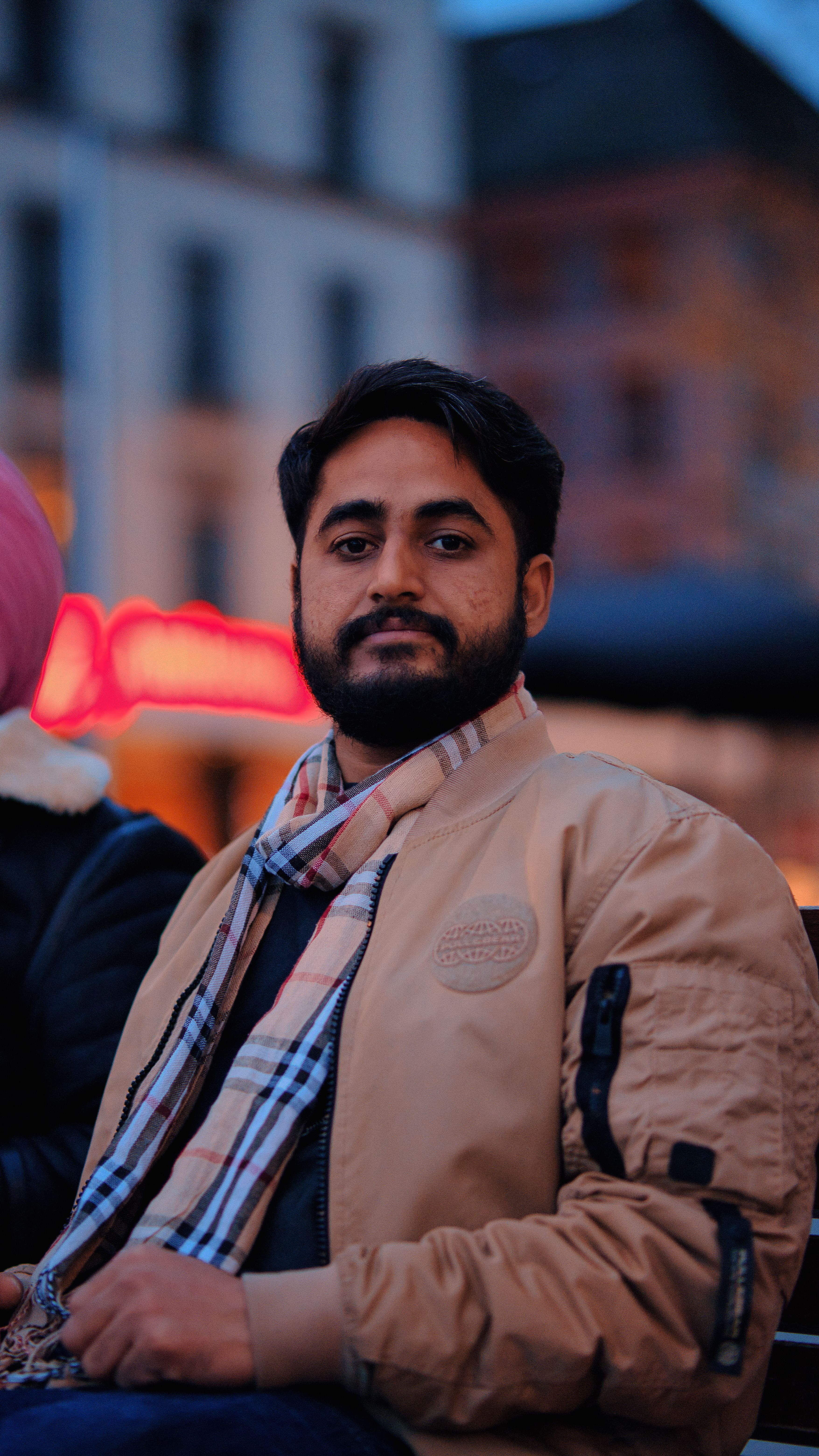 A portrait of a man with a beard and mustache, wearing a jacket and shawl, in an urban setting during twilight.