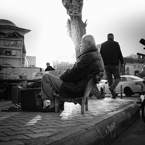 Candid Shot of an Elderly Man Sitting on a Chair on a Sidewalk and Selling Items 