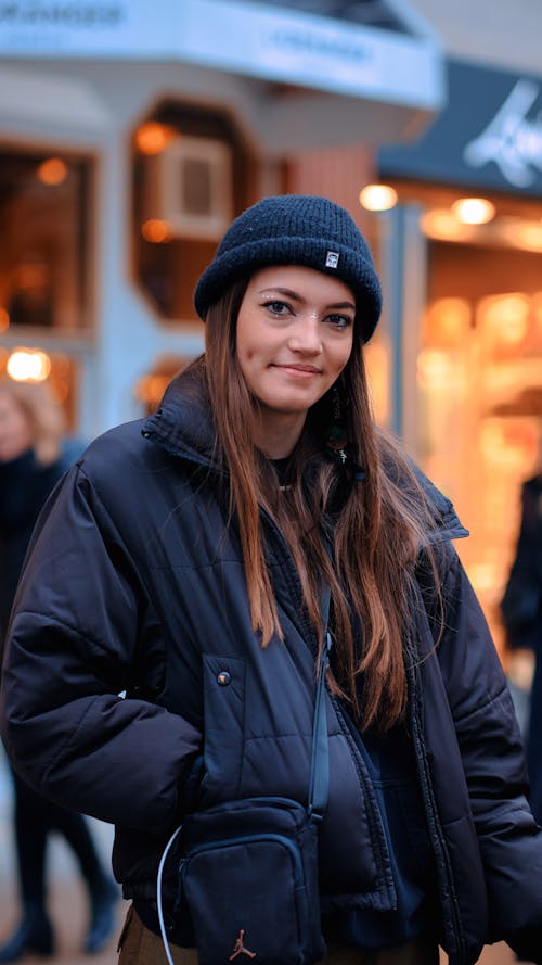 Young Woman in a Jacket and Hat Standing in City and Smiling 
