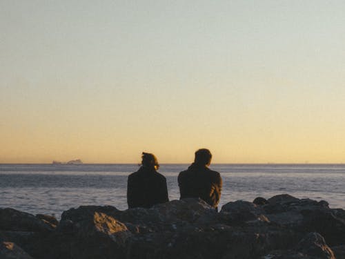 Back View of Two People Sitting on a Rocky Shore and Looking at the Sea at Sunset