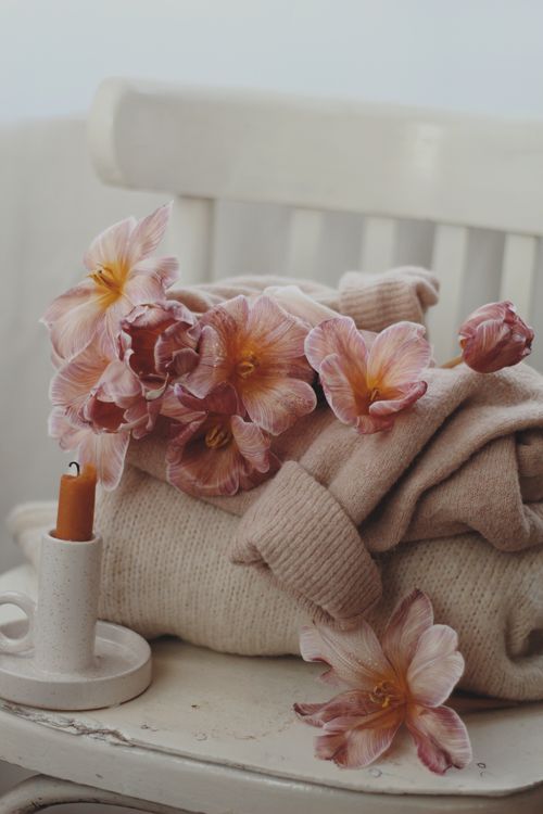 Free A stack of beige knit sweaters with pink flowers and a candle on a chair, creating a cozy setting. Stock Photo