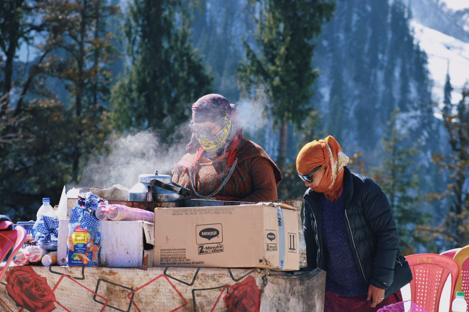 Two women cook outdoors in snowy Manali, India, amidst mountains and steam.