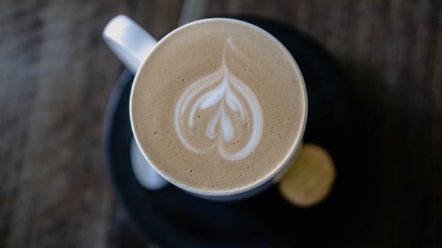 Top-down view of a cappuccino with intricate froth art in a white cup on a dark table.