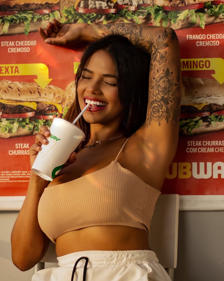 Young Woman With Tattoos Smiling And Drinking Soda From A Disposable Cup 