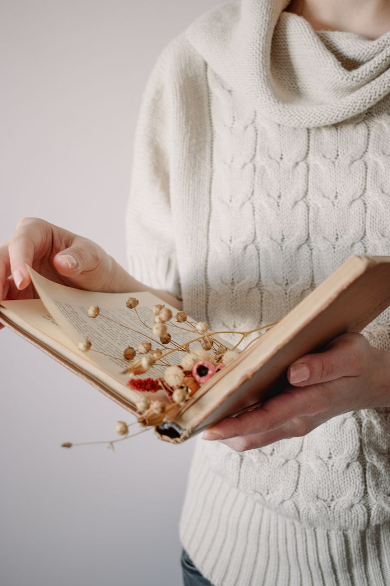A woman wearing a white sweater holds an open vintage book with dried flowers between the pages. A woman wearing a white sweater holds an open vintage book with dried flowers between the pages.