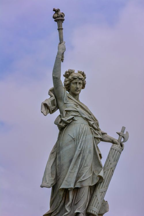 Free Elegant marble statue depicting a woman holding a torch and shield against a cloudy sky. Stock Photo