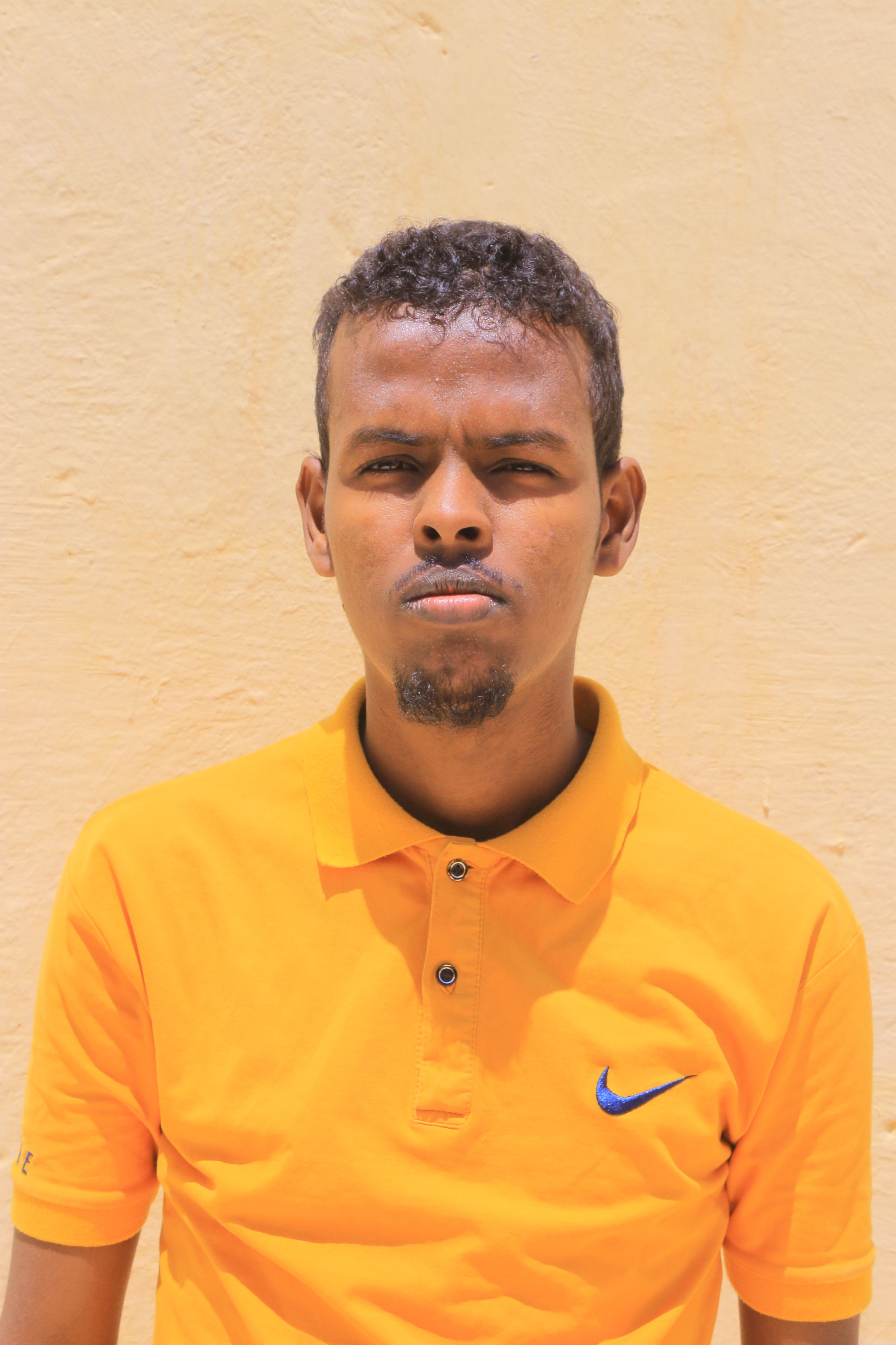 Portrait of a young man wearing a bright orange polo shirt, standing against a textured wall in Galkayo, Somalia.