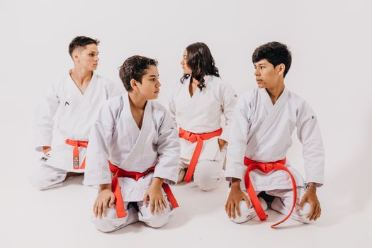 Group of teenagers in karate uniforms with red belts sitting in a studio setting.