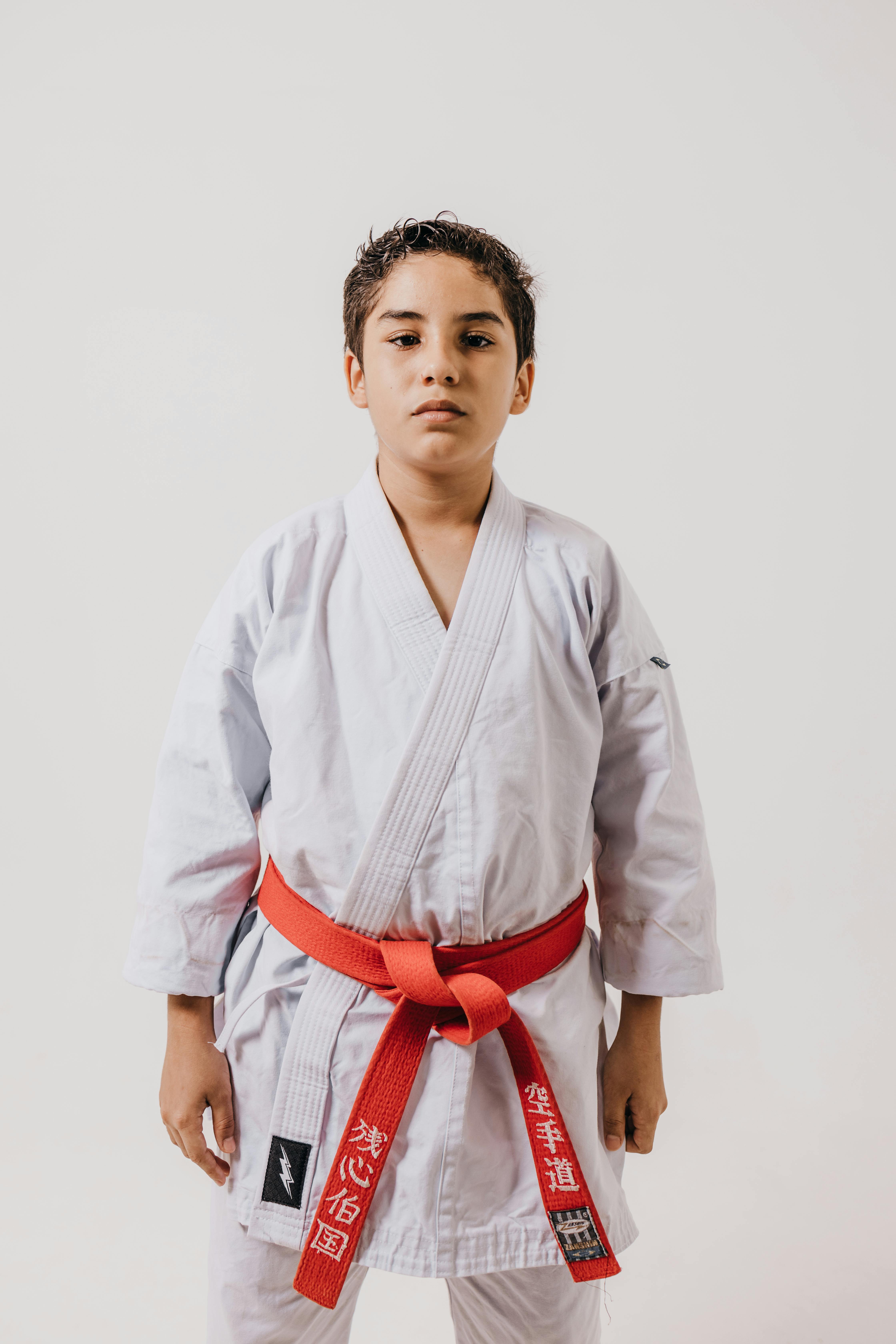 A young martial artist in a white kimono with a red belt poses confidently in a studio setting.