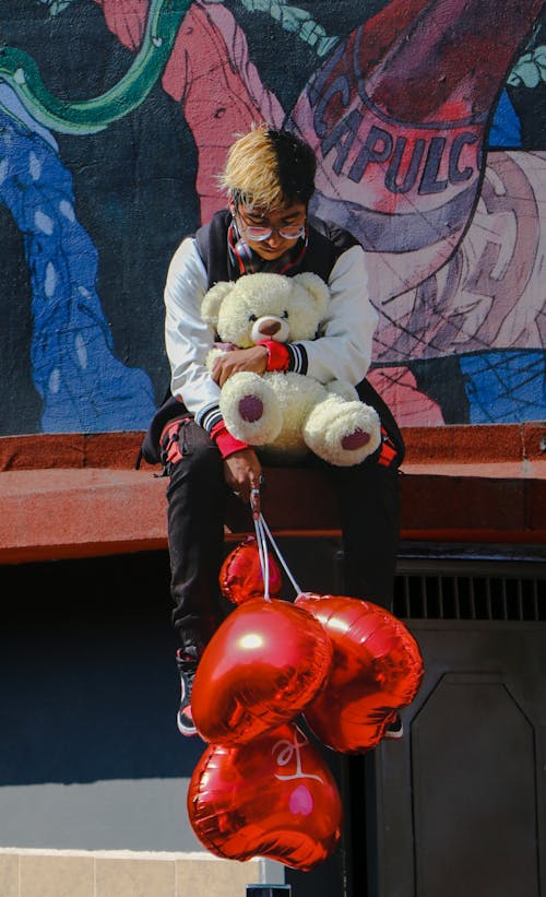 A Young Man Sitting Alone and Holding Heart Shaped Balloons and a Teddy Bear 