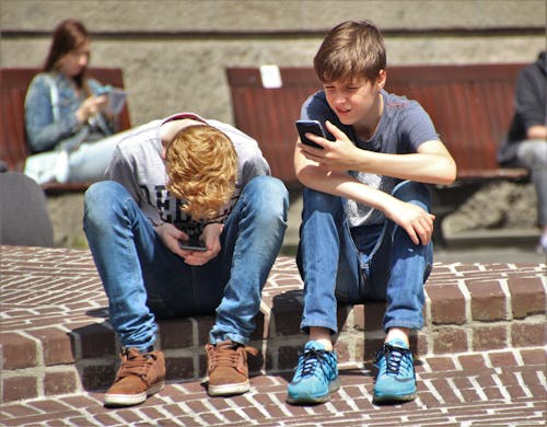 Free 2個男孩坐在棕色地板上，一邊使用他們的智能手機，一邊白天白天坐在長凳上使用智能手機的女人 Stock Photo